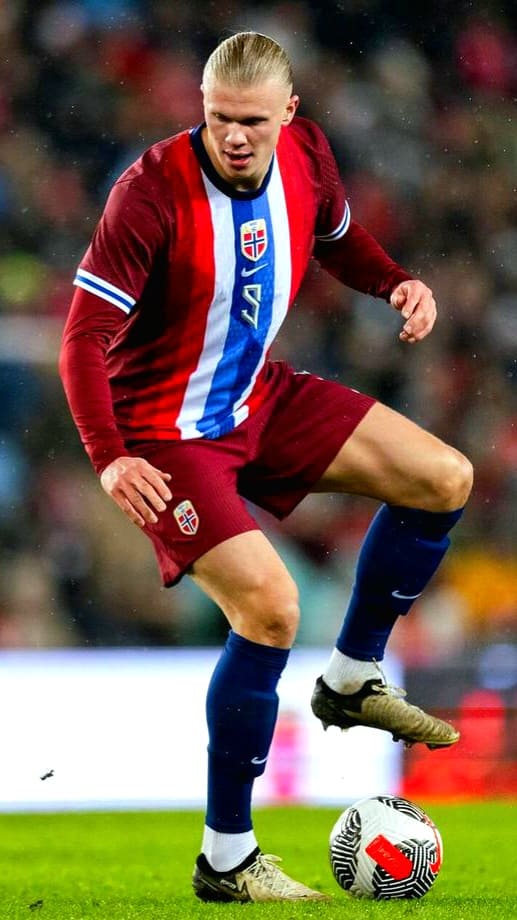 A male soccer player in a red and blue Norway national team uniform controls a soccer ball on the field during a match, as seen on Ecuador IPTV. The stadium lights and crowd are visible in the blurred background.
