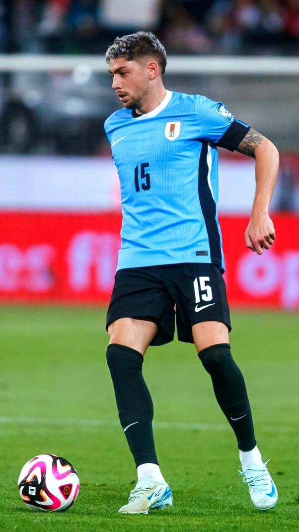 A soccer player in a light blue jersey with the number 15 controls the ball on a field during a match, as fans watch live via Ecuador IPTV. The player sports black shorts, black socks, and white shoes.