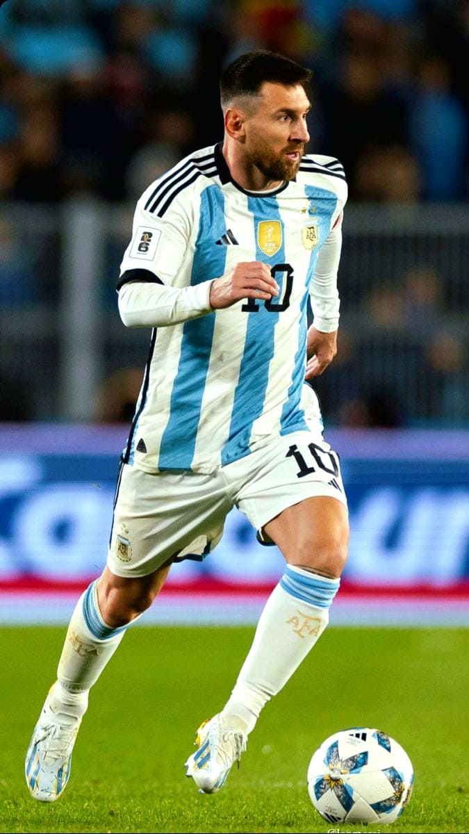 A soccer player in an Argentina jersey, number 10, runs on the field while controlling a soccer ball during a match, as spectators and an Ecuador IPTV banner blur in the background.